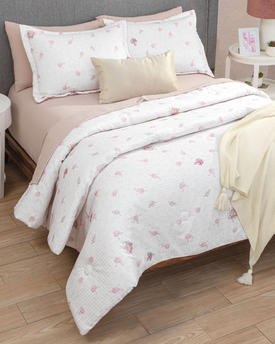 A neatly made bed dressed in the Juego de Edredón Luxury Jardín by Intima Hogar, featuring a white floral comforter, pink sheet, matching pillows, a beige accent pillow, and a cream throw, invites you into this serene recámara. The room boasts wood floors and a nightstand with a photo frame and lamp—everything you need for un descanso reparador.