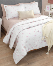 A neatly made bed dressed in the Juego de Edredón Luxury Jardín by Intima Hogar, featuring a white floral comforter, pink sheet, matching pillows, a beige accent pillow, and a cream throw, invites you into this serene recámara. The room boasts wood floors and a nightstand with a photo frame and lamp—everything you need for un descanso reparador.