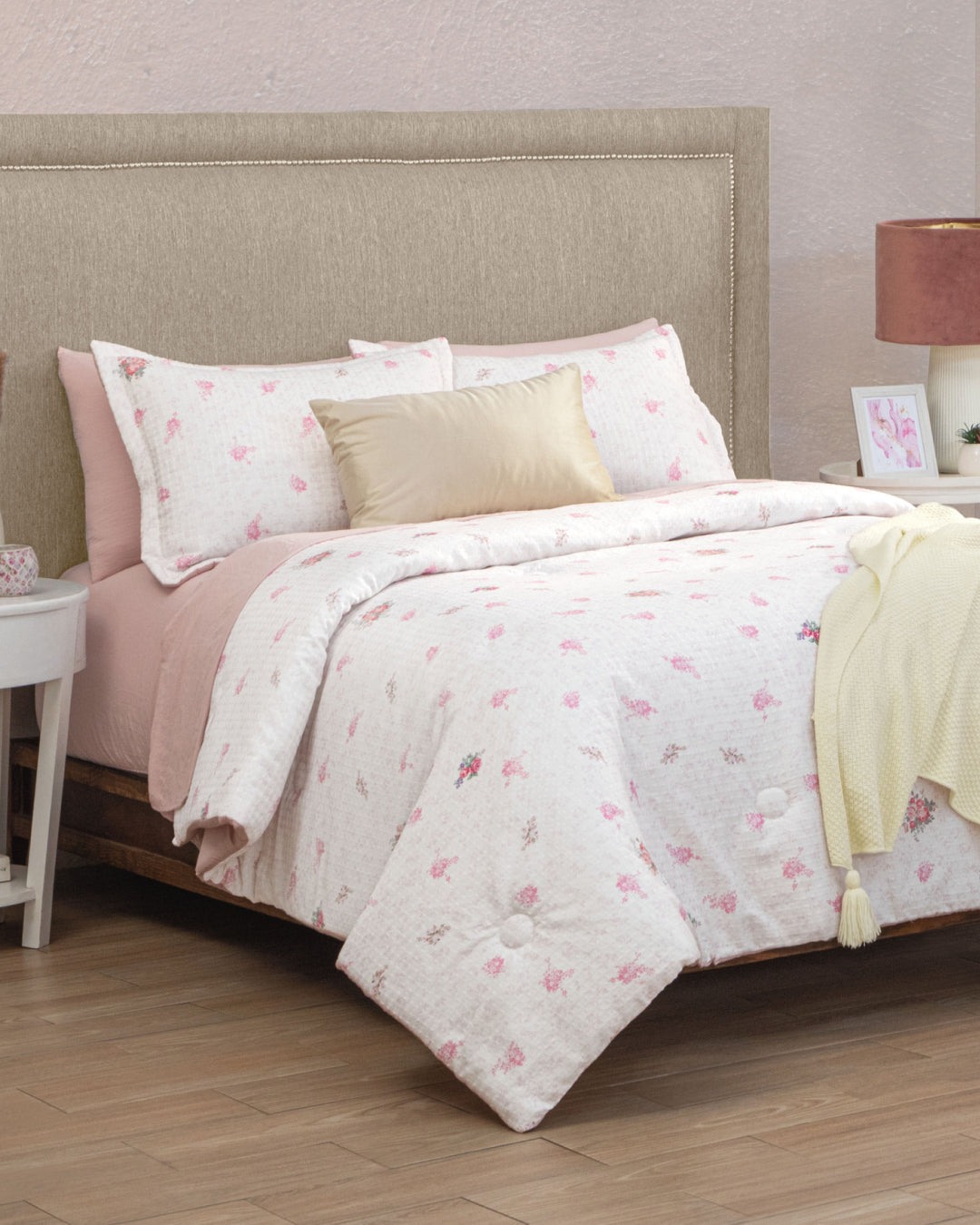 A neatly made bed with a beige headboard, dressed in the Juego de Edredón Luxury Jardín by Intima Hogar featuring white and pink floral patterns, paired with a gold pillow and a yellow knit blanket. The bed is flanked by white nightstands topped with a lamp and decorative accents, all set on a wooden floor—creating the perfect recámara for un descanso reparador.