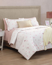 A neatly made bed with a beige headboard, dressed in the Juego de Edredón Luxury Jardín by Intima Hogar featuring white and pink floral patterns, paired with a gold pillow and a yellow knit blanket. The bed is flanked by white nightstands topped with a lamp and decorative accents, all set on a wooden floor—creating the perfect recámara for un descanso reparador.