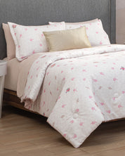 Transforming the sentence using your product data: A neatly made bed with the Juego de Edredón Luxury Jardín by Intima Hogar, featuring a white edredón and pillow shams adorned with delicate pink floral patterns, complemented by light pink sheets and a gold decorative pillow, set against a gray upholstered headboard, invites you into the recámara for un descanso reparador.