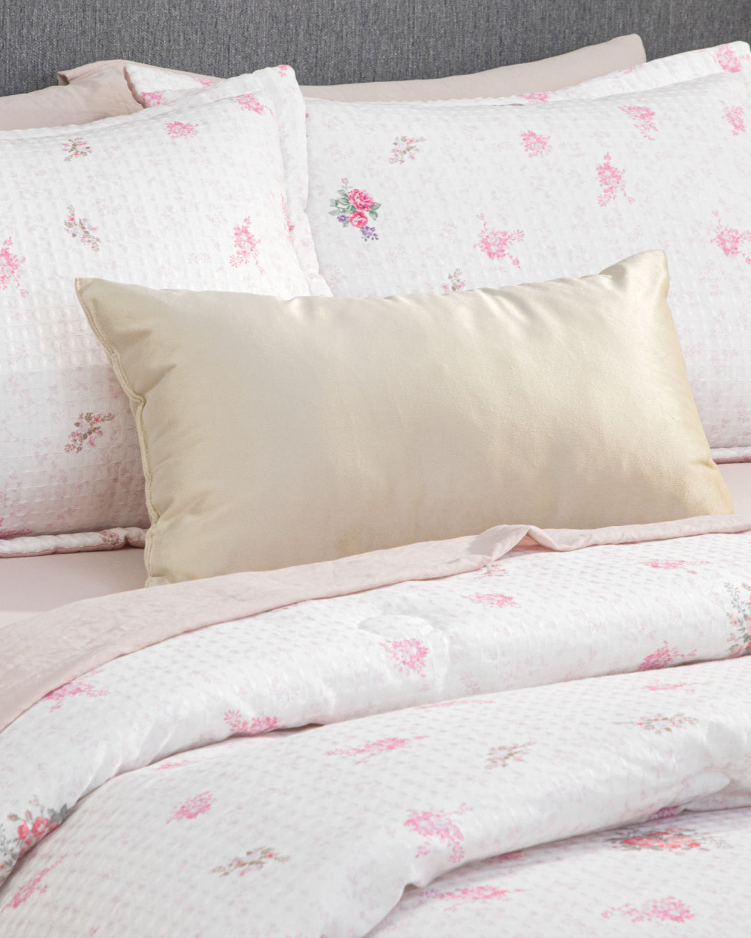 A neatly made bed featuring the Intima Hogar Juego de Edredón Luxury Jardín, with its white and pink floral design, a soft light pink sheet, and a shiny beige accent pillow artfully placed in front of matching floral pillows—all set against a gray upholstered headboard—creates the perfect atmosphere for un descanso reparador in your recámara.
