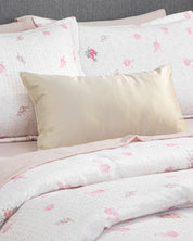 A neatly made bed featuring the Intima Hogar Juego de Edredón Luxury Jardín, with its white and pink floral design, a soft light pink sheet, and a shiny beige accent pillow artfully placed in front of matching floral pillows—all set against a gray upholstered headboard—creates the perfect atmosphere for un descanso reparador in your recámara.