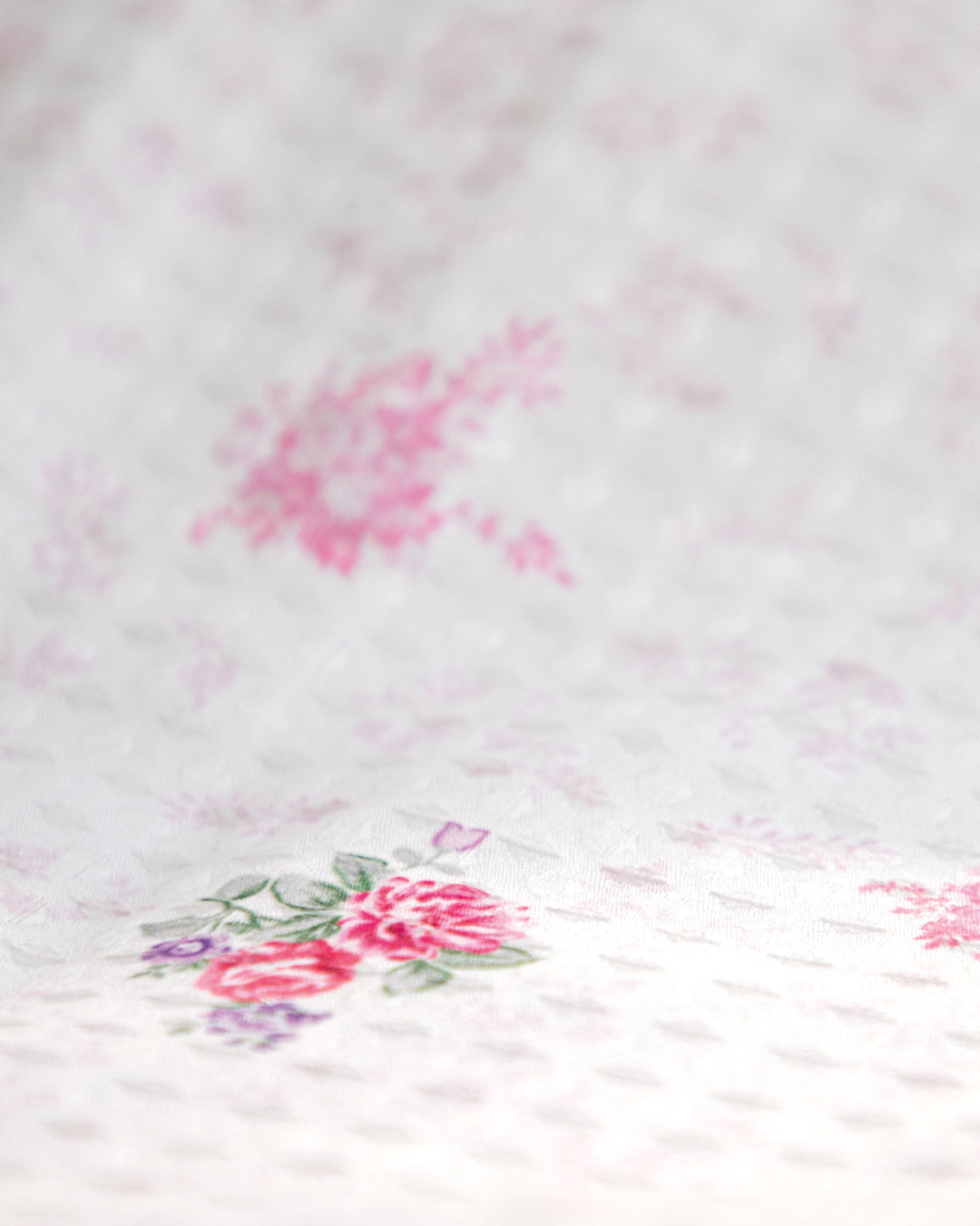 Un primer plano del Juego de Edredón Luxury Jardín de Intima Hogar muestra su suave tela blanca con un delicado estampado floral en tonos rosados, realzado por hojas verdes y pequeños detalles en morado. La textura ligeramente elevada crea un sutil efecto acolchado, perfecto para agregar encanto y comodidad a cualquier recámara, creando el ambiente ideal para un descanso reparador.