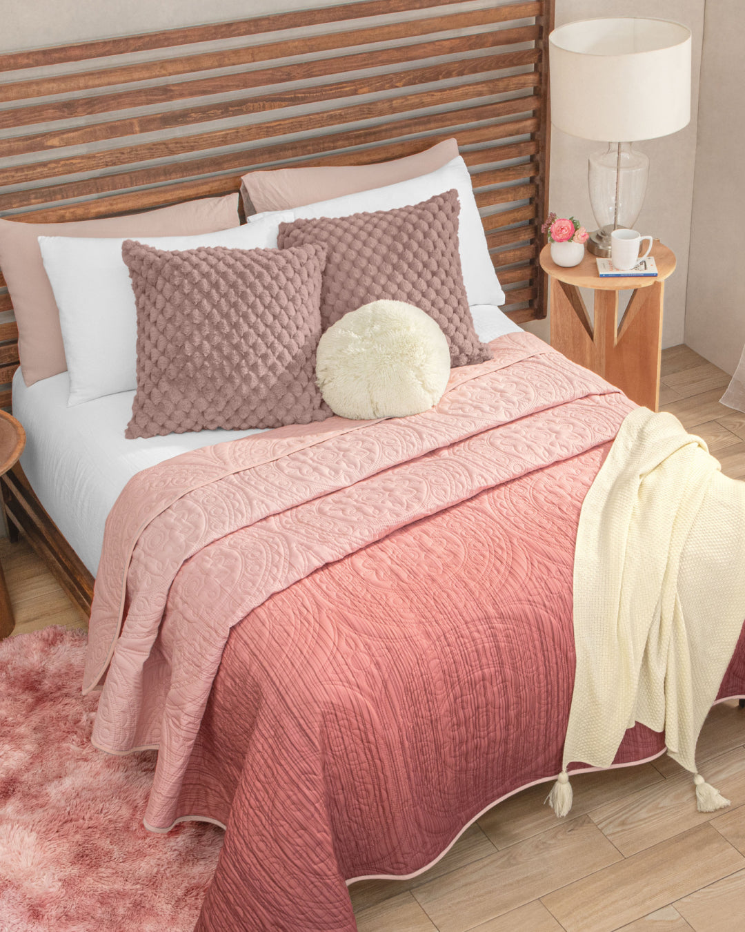A neatly made bed features the Juego de Edredón Ligero Carisma by Intima Hogar in a pink ombre pattern, complemented by textured pillows, a round cream cushion, a cream throw, and a wooden headboard with textura capitonada. Next to the bed, a wooden nightstand displays a lamp, books, flowers, and a mug. A pink rug lies on the floor.