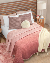 A neatly made bed features the Juego de Edredón Ligero Carisma by Intima Hogar in a pink ombre pattern, complemented by textured pillows, a round cream cushion, a cream throw, and a wooden headboard with textura capitonada. Next to the bed, a wooden nightstand displays a lamp, books, flowers, and a mug. A pink rug lies on the floor.