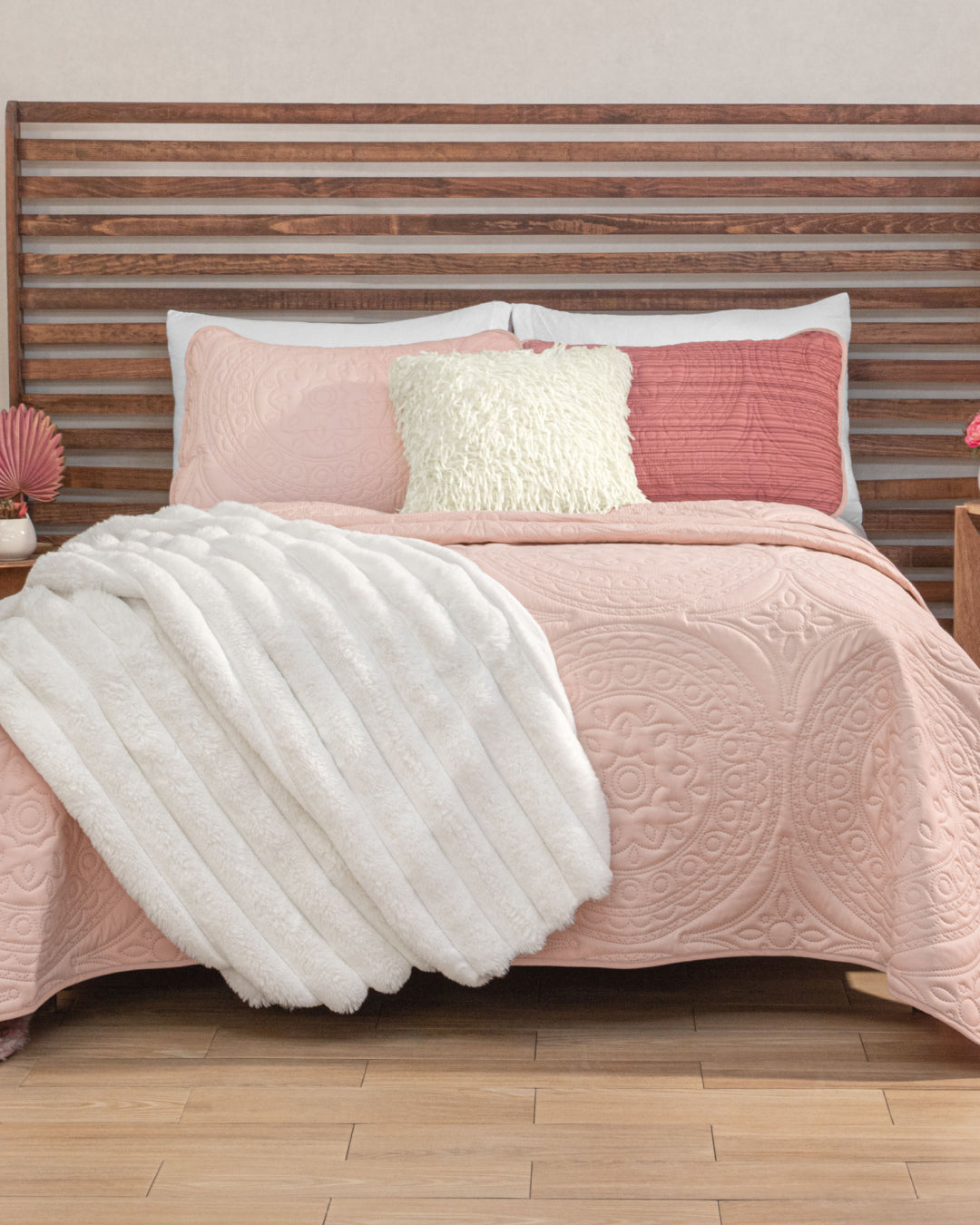 A neatly made bed features the Juego de Edredón Ligero Carisma by Intima Hogar in pink textura capitonada, complemented by a white fluffy blanket and cream and pink pillows with fundas decorativas. The wooden slat headboard pairs with two nightstands adorned with small decorative items on either side.