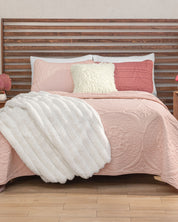 A neatly made bed features the Juego de Edredón Ligero Carisma by Intima Hogar in pink textura capitonada, complemented by a white fluffy blanket and cream and pink pillows with fundas decorativas. The wooden slat headboard pairs with two nightstands adorned with small decorative items on either side.