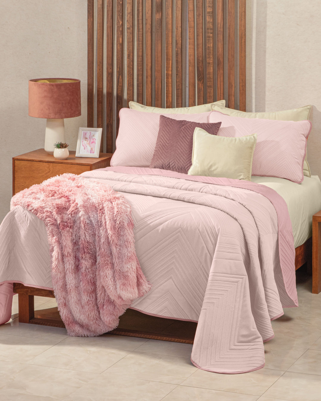 A cozy recámara features a wooden bed dressed with the Juego de Edredón Ligero Mauve by Intima Hogar, complemented by a textured pink throw blanket and pastel decorative pillow covers. A nightstand holds a pink lamp and framed photo. The tiled floor enhances the room’s soft and inviting decor.