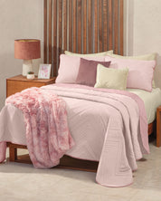 A cozy recámara features a wooden bed dressed with the Juego de Edredón Ligero Mauve by Intima Hogar, complemented by a textured pink throw blanket and pastel decorative pillow covers. A nightstand holds a pink lamp and framed photo. The tiled floor enhances the room’s soft and inviting decor.