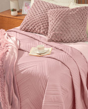 A neatly made bed in the recámara showcases the Juego de Edredón Ligero Mauve by Intima Hogar, featuring textured pink fundas decorativas, a quilted pink edredón, and a matching pink throw. A tray with a cup and saucer rests on the bed, while a wooden nightstand with a photo and lamp sits nearby.