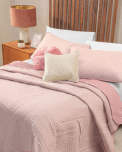 The bed in this recámara is neatly made with the Juego de Edredón Ligero Mauve from Intima Hogar, featuring pink geometric bedding and a matching edredón. It is complemented by a white pillow, a fuzzy pink pillow, and cream fundas decorativas. Behind the bed stands a wooden headboard, while a nightstand with a lamp, framed picture, and small plant sits beside it.