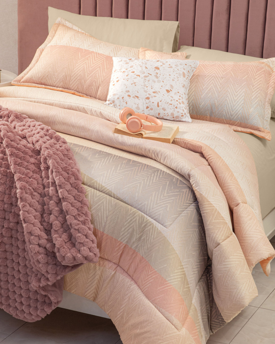 A neatly made bed features the Juego de Edredón Suave by Intima Hogar in beige and peach tones, complemented by a pink textured throw, decorative pillows, an open book, and pink headphones on top. The mauve-upholstered headboard enhances the inviting atmosphere for confort diario and descanso reparador.
