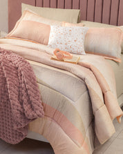 A neatly made bed features the Juego de Edredón Suave by Intima Hogar in beige and peach tones, complemented by a pink textured throw, decorative pillows, an open book, and pink headphones on top. The mauve-upholstered headboard enhances the inviting atmosphere for confort diario and descanso reparador.