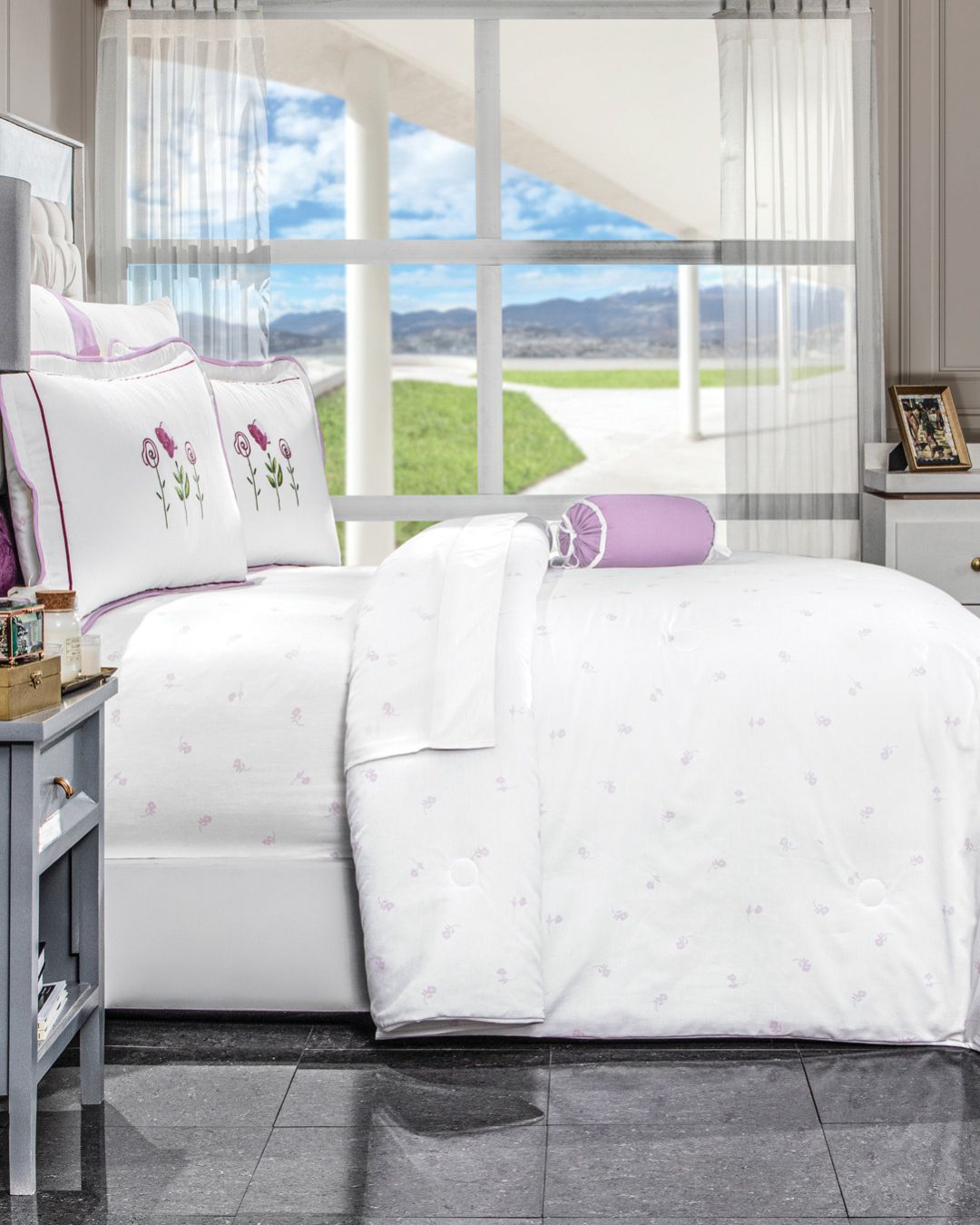 A neatly made bed features the Juego de Edredón Amore by Intima Hogar and pillows with purple flower embroidery, set before a large window with sheer curtains, showcasing mountain and green grass views.