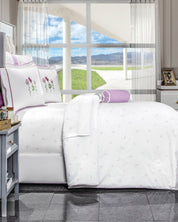 A neatly made bed features the Juego de Edredón Amore by Intima Hogar and pillows with purple flower embroidery, set before a large window with sheer curtains, showcasing mountain and green grass views.