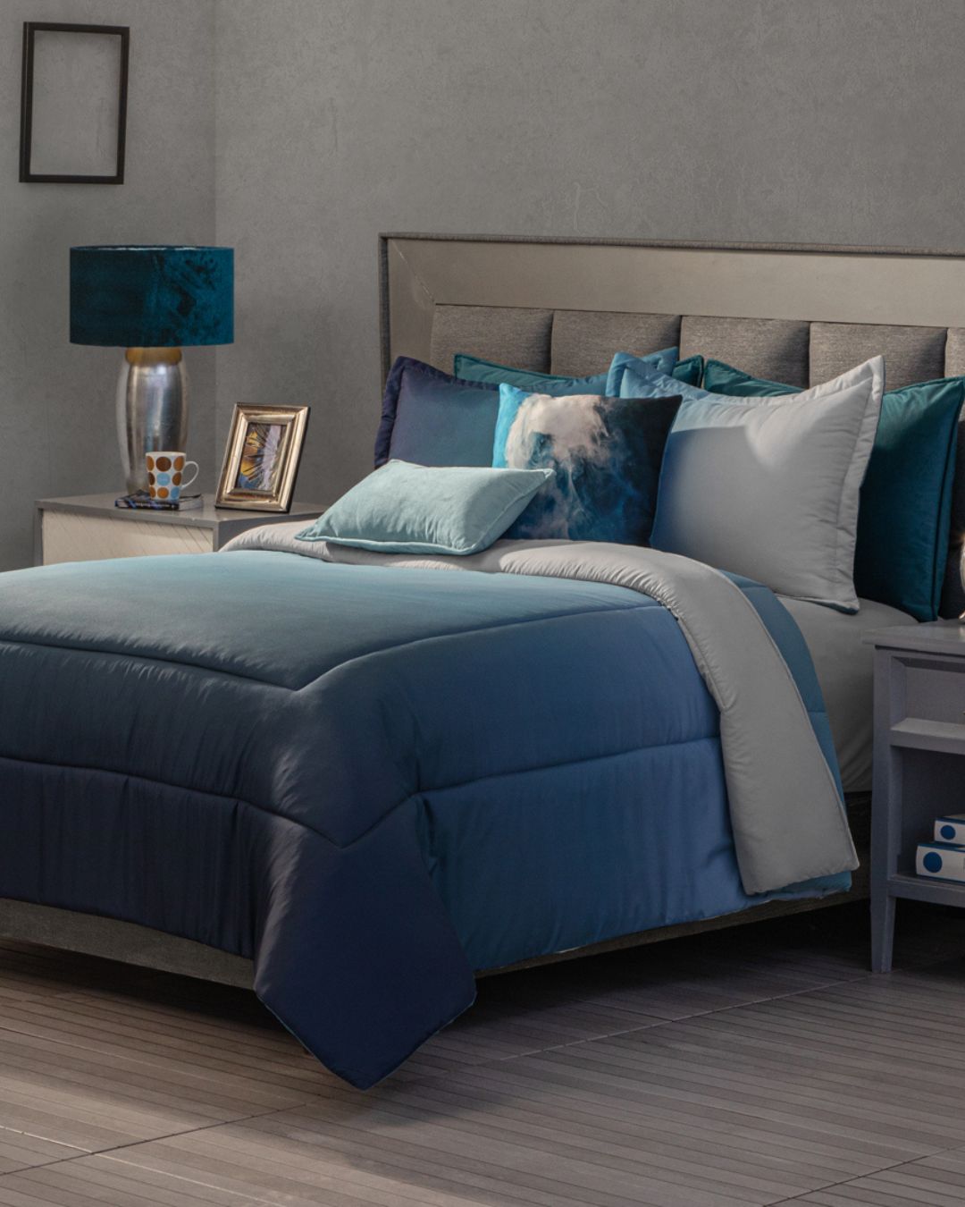 A neatly made bed features the Juego de Edredón Blue by Intima Hogar, accented with decorative pillows. A nightstand holds a lamp, framed photo, and cup, while gray wall decor and a small framed picture complete the look.