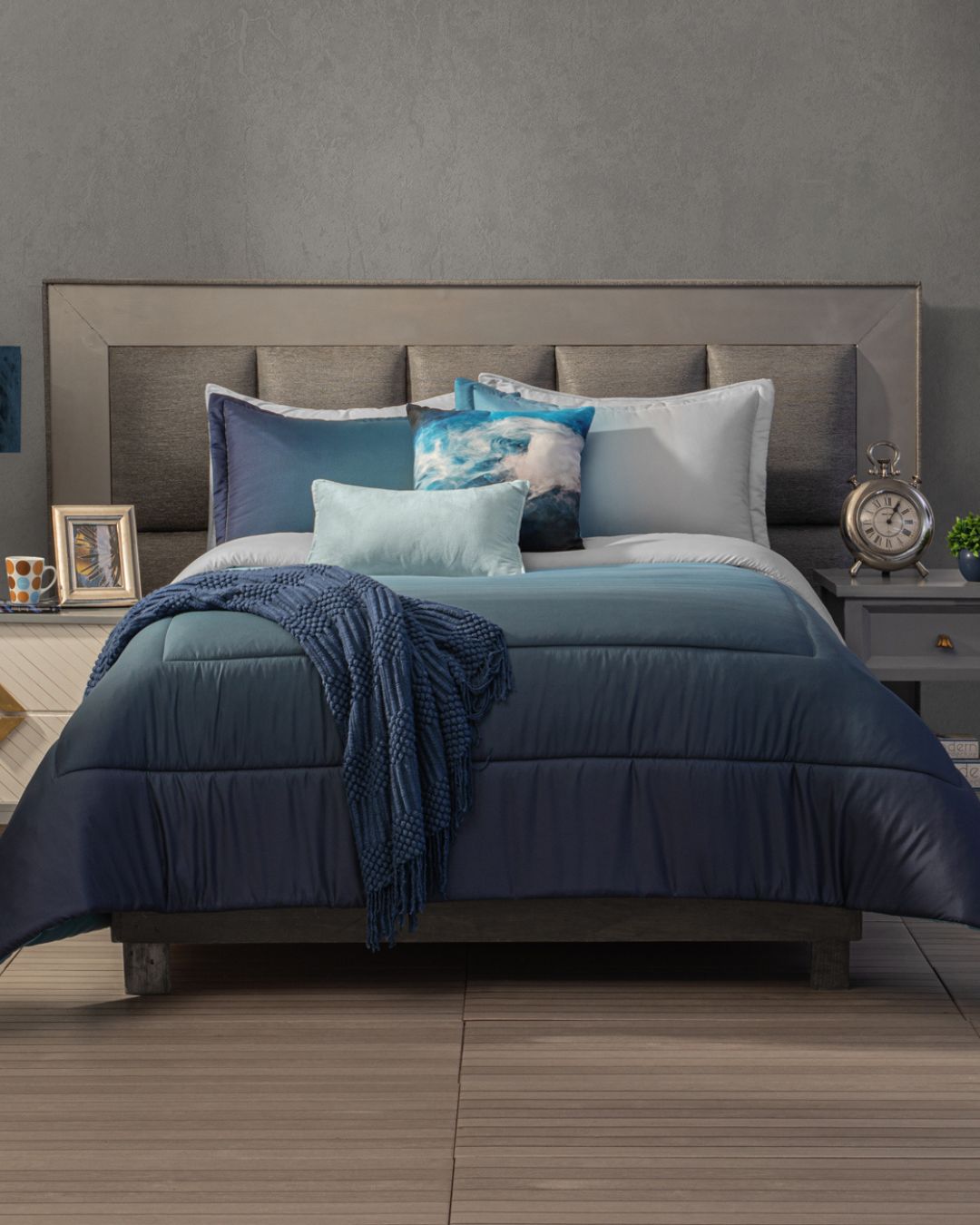 The bed features the Intima Hogar Juego de Edredón Blue with blue and white bedding, decorative pillows, and a blue throw. It’s centered against a gray wall, with a nightstand holding a clock and lamp on the right side.
