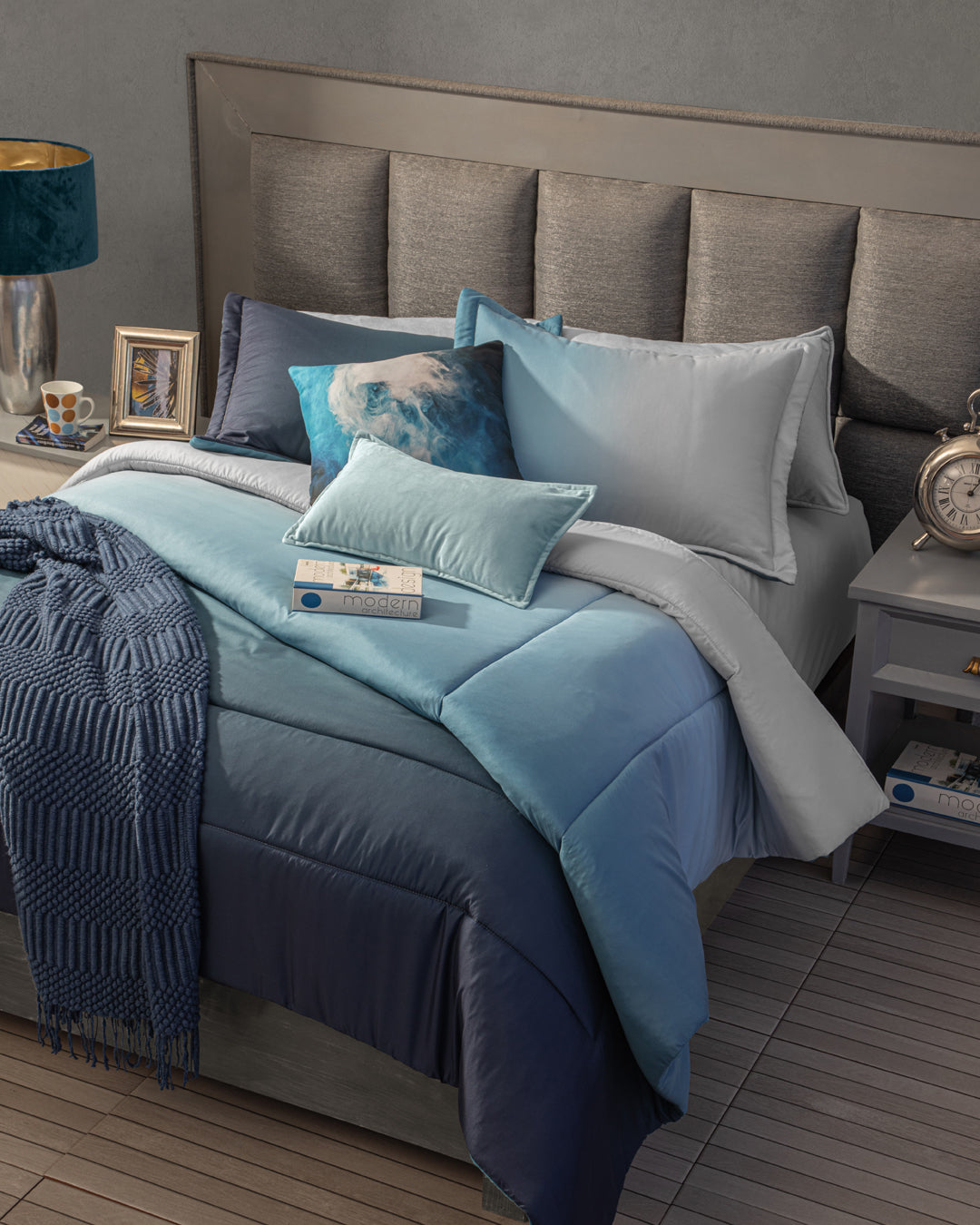 A neatly made bed with the Juego de Edredón Blue by Intima Hogar, several pillows, and a textured throw sits beside a gray nightstand with a lamp, clock, photo frame, and cup for a cozy bedroom scene.