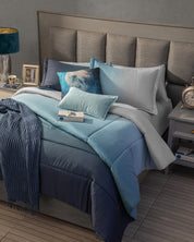 A neatly made bed with the Juego de Edredón Blue by Intima Hogar, several pillows, and a textured throw sits beside a gray nightstand with a lamp, clock, photo frame, and cup for a cozy bedroom scene.