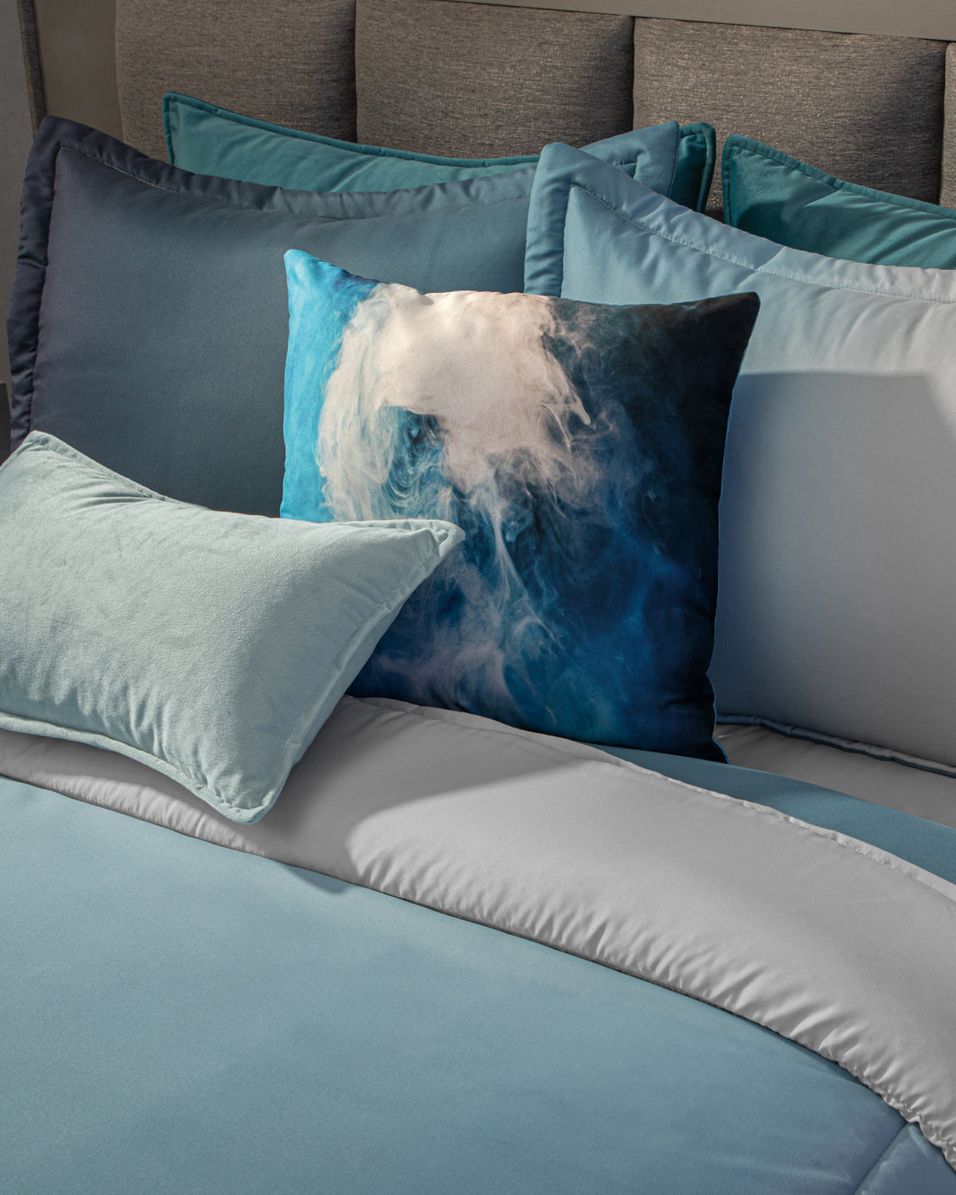 A neatly made bed showcases the Juego de Edredón Blue by Intima Hogar, featuring blue and gray bedding and several pillows, including a square pillow with a blue, black, and white abstract design at the center.