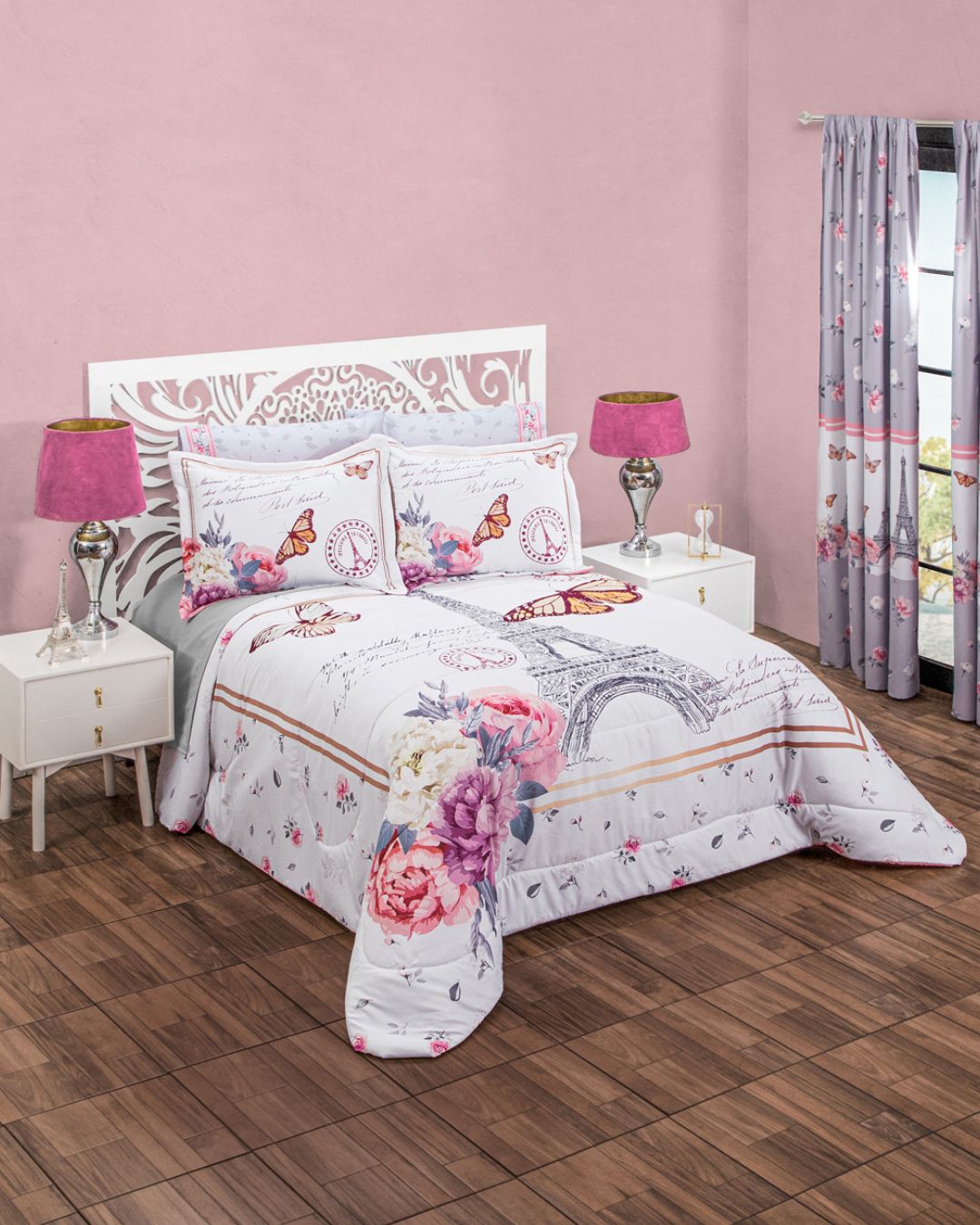 A bedroom featuring the Juego de Edredón Súper Suave Camil by Intima Hogar, with a white ornate bed frame, butterfly and floral bedding, pink lamps on white nightstands, pink walls, wood floor, and gray floral curtains.