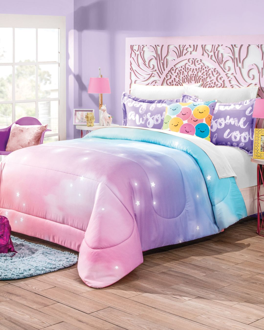 A bed with the Juego de Edredón Dreams by Intima Hogar features a pastel ombre design in pink, purple, and blue with white stars. Colorful smiley face pillows and “awesome” text, plus lavender walls and a floral headboard create un diseño mágico.
