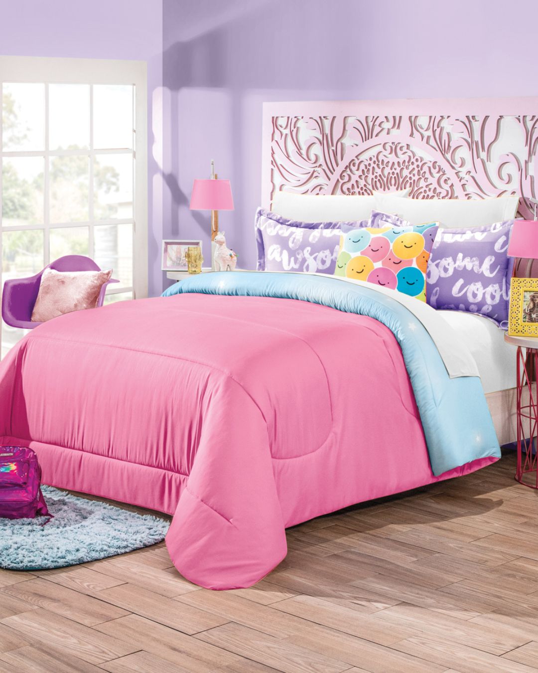 A neatly made bed with the Juego de Edredón Dreams by Intima Hogar in pink, light blue sheets, and colorful pillows sits in a lavender bedroom with sunlight streaming in. A purple backpack and chair complete the diseño mágico near the bed.