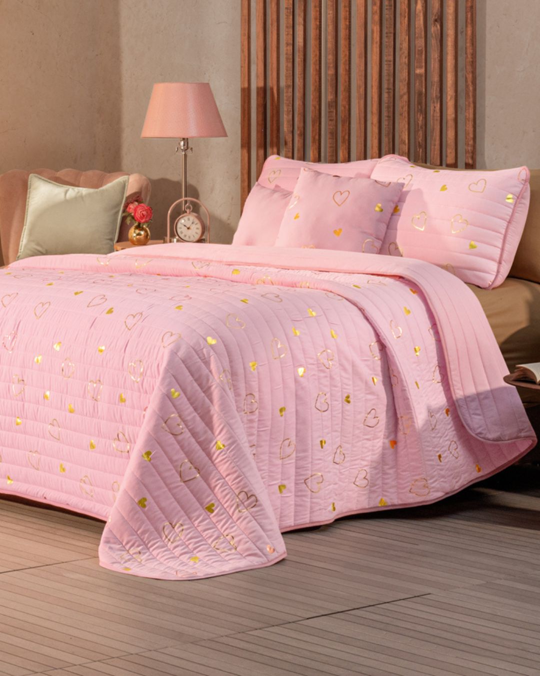 A cozy, modern bedroom features the Intima Hogar Juego de Edredón Dulce in pink with heart-patterned pillows and decorative cushions, beside a nightstand holding a lamp, alarm clock, and cushion.