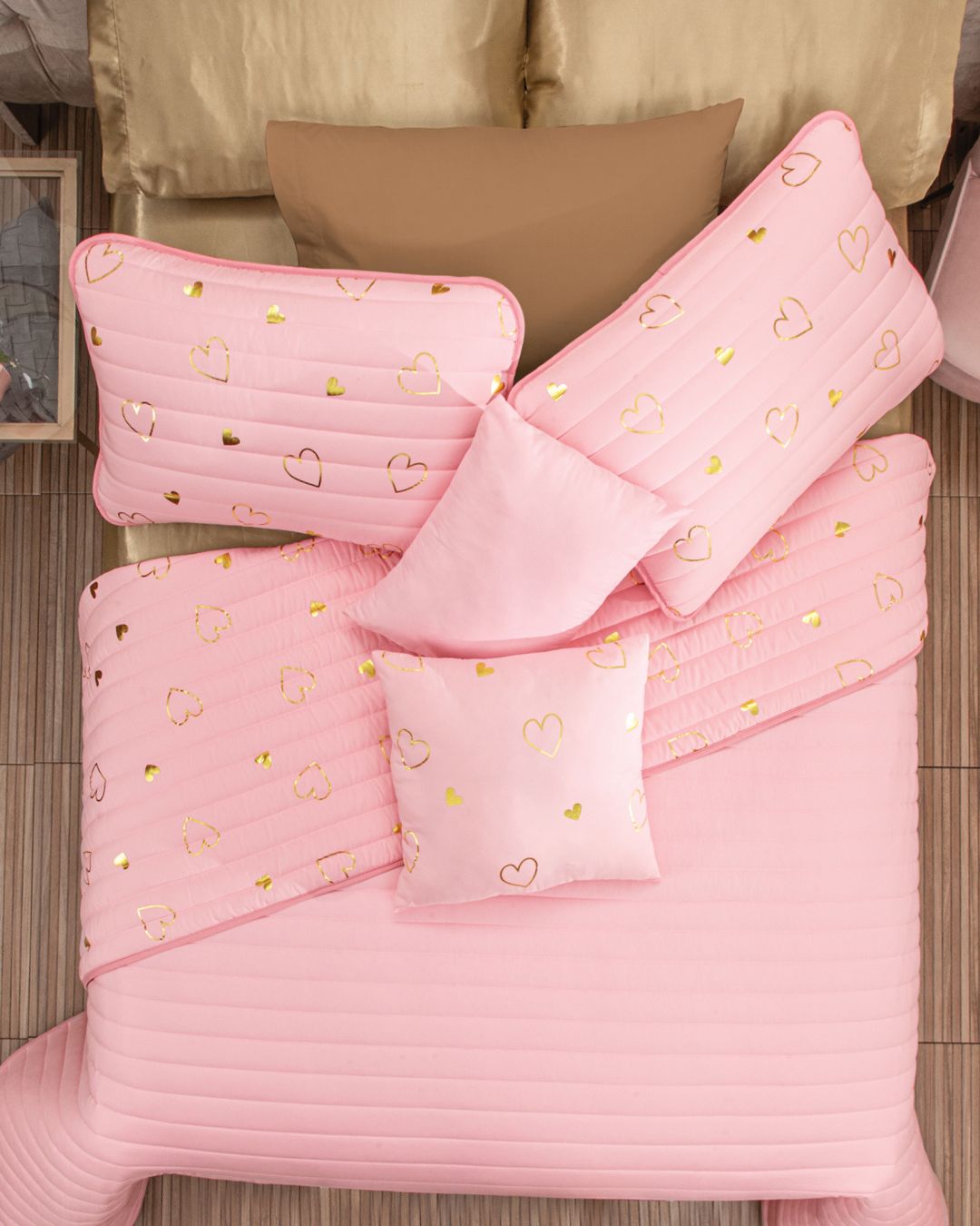 A neatly made bed features the Intima Hogar Juego de Edredón Dulce in pink with gold heart pillows and tan fundas decorativas, set on a light wooden floor.