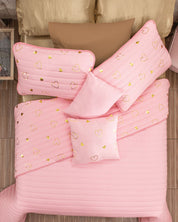 A neatly made bed features the Intima Hogar Juego de Edredón Dulce in pink with gold heart pillows and tan fundas decorativas, set on a light wooden floor.