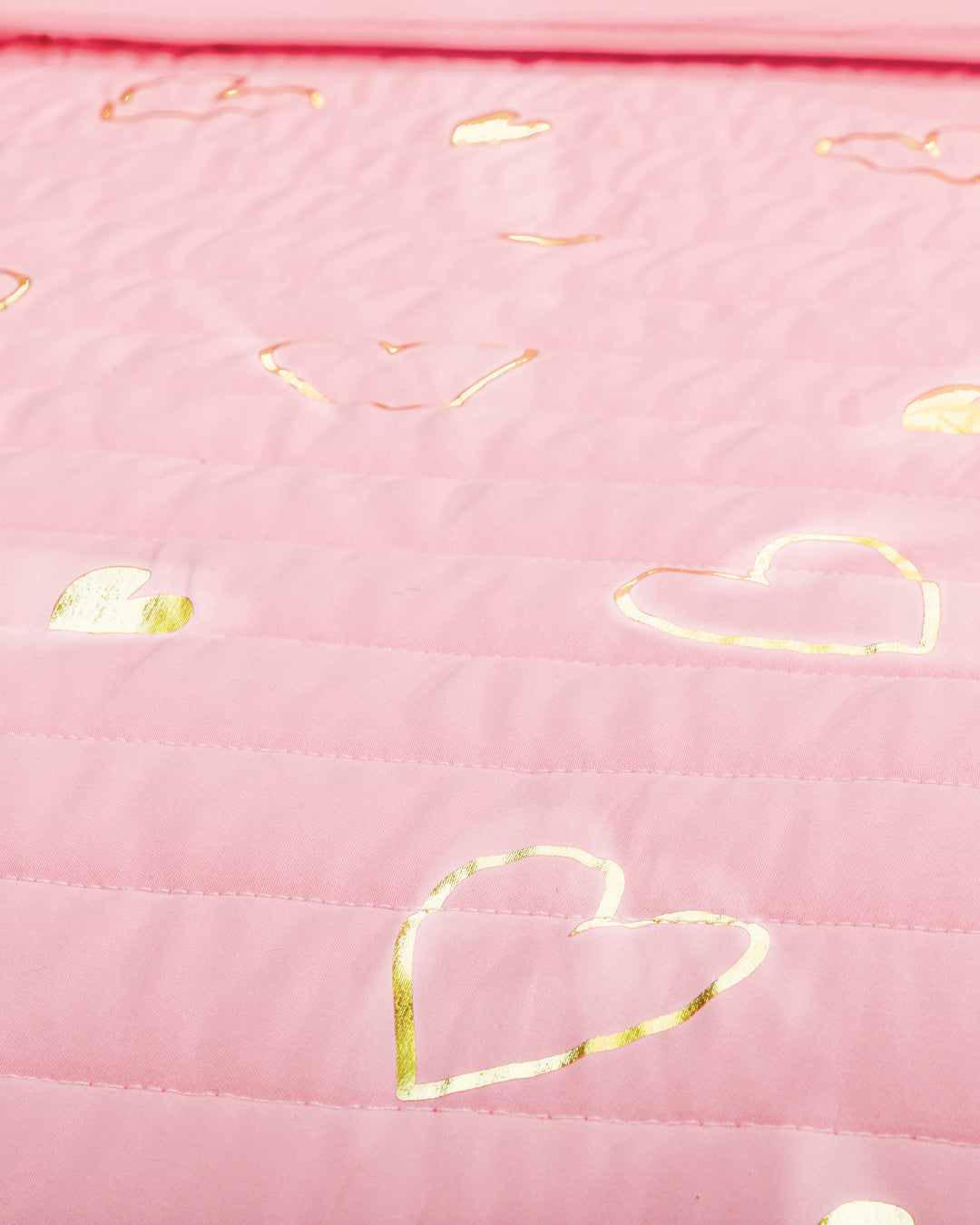 A close-up of the Juego de Edredón Dulce by Intima Hogar shows pink quilted fabric with shiny gold heart outlines scattered across the surface.