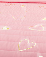 A close-up of the Juego de Edredón Dulce by Intima Hogar shows pink quilted fabric with shiny gold heart outlines scattered across the surface.