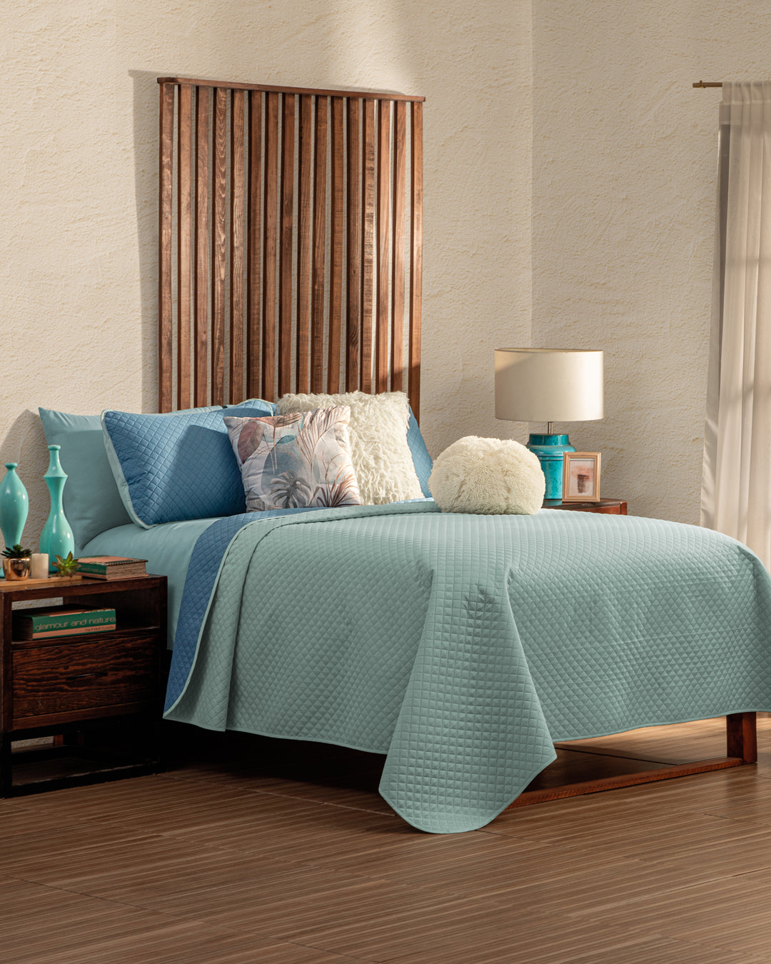 A neatly made bed features the Juego de Edredón Edén by Intima Hogar in blue and white, topped with decorative pillows and a wooden headboard. A side table displays turquoise vases, a lamp, and books beside a bright window with white curtains.