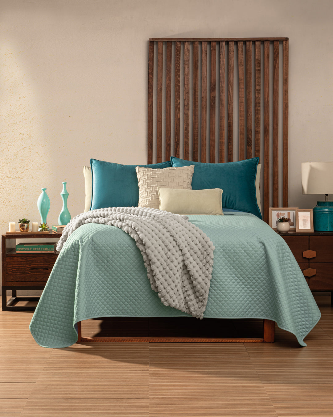 A neatly made bed featuring the Intima Hogar Juego de Edredón Edén, assorted pillows, and a textured gray blanket, flanked by wooden nightstands with decor items, set against a beige wall with a vertical wood panel headboard.