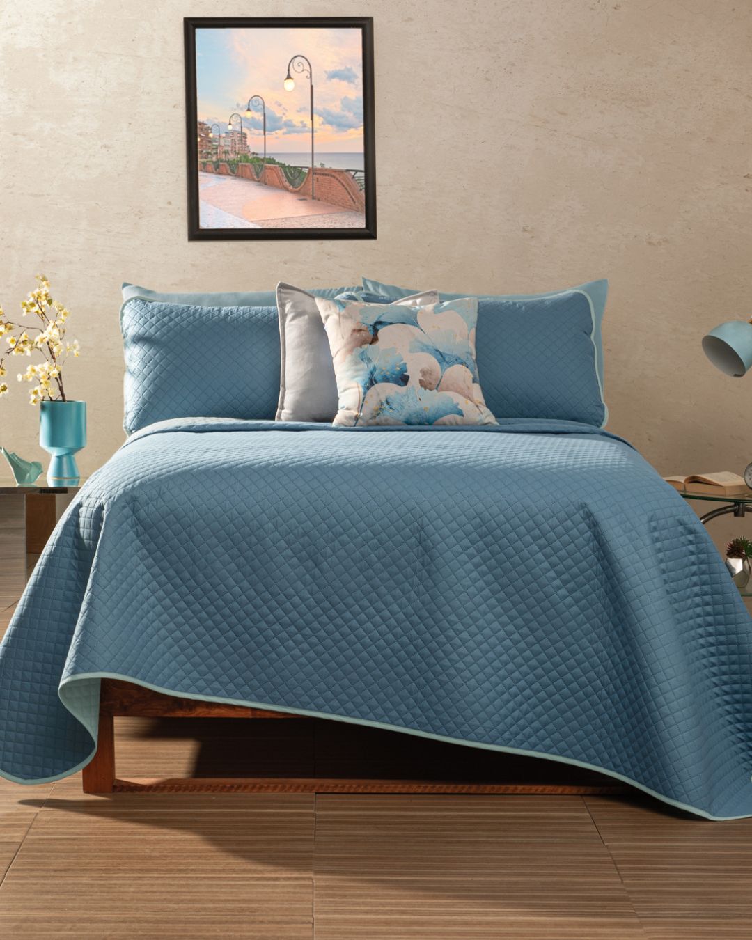 A neatly made bed with the Juego de Edredón Edén de Intima Hogar and decorative shams stands against a beige wall. Above hangs a seaside painting, while yellow flowers in a vase and a lamp decorate the bedside tables.