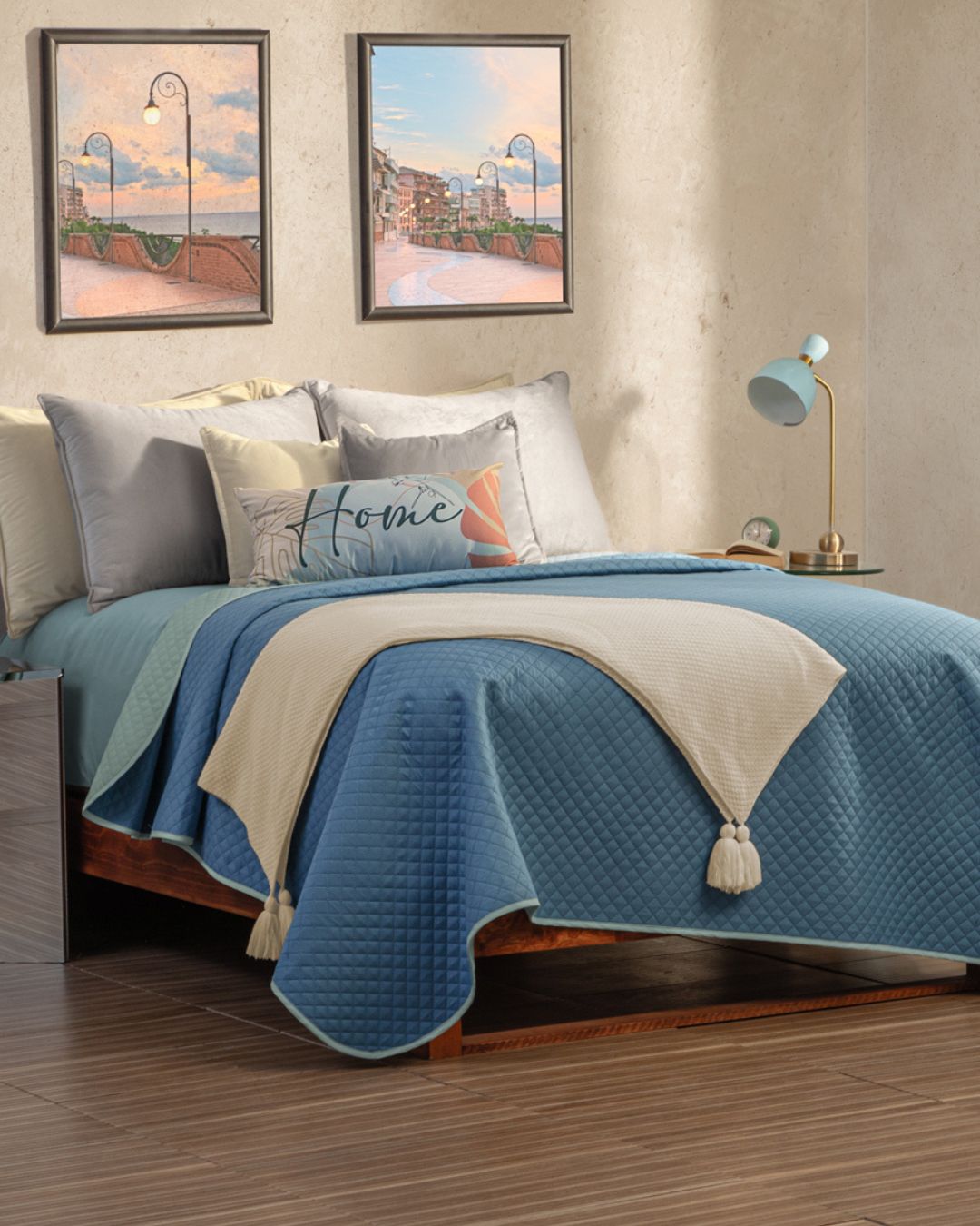 A neatly made bed with the Juego de Edredón Edén by Intima Hogar in blue and white, decorative pillows (one reads "Home"), a blue lamp on the bedside table, and two scenic street paintings framed above the bed.