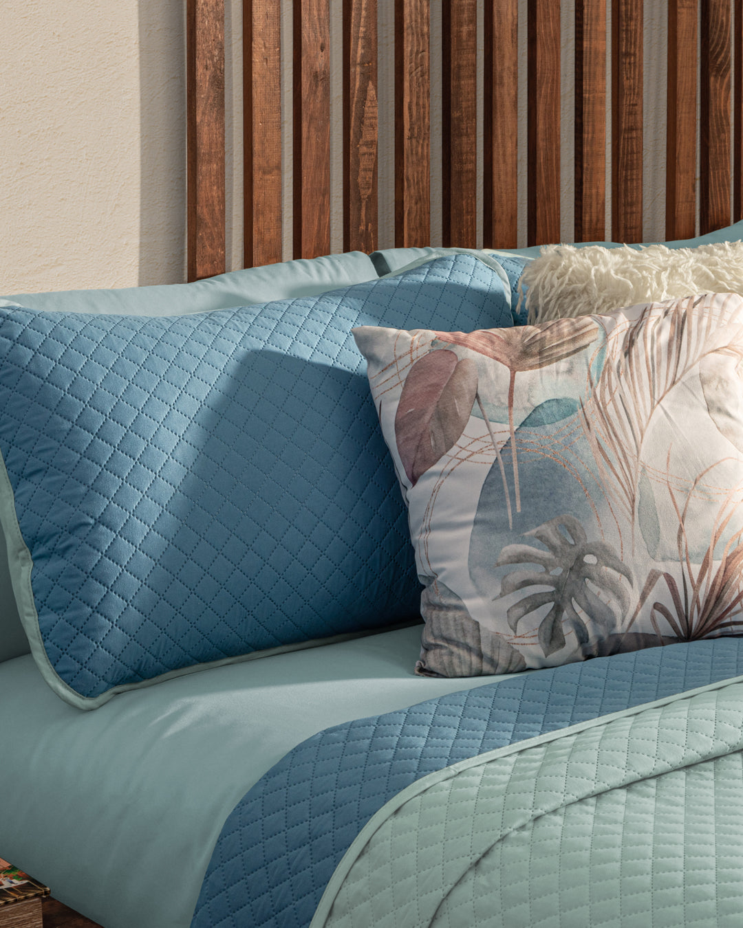 A neatly made bed features the Juego de Edredón Edén bedding set by Intima Hogar, with light blue quilted covers, matching pillows, and a pastel botanical decorative pillow against a wooden slatted headboard.