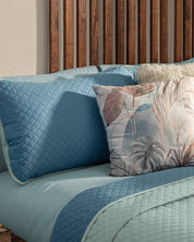 A neatly made bed features the Juego de Edredón Edén bedding set by Intima Hogar, with light blue quilted covers, matching pillows, and a pastel botanical decorative pillow against a wooden slatted headboard.