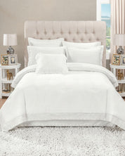 A neatly made bed with the Juego de Edredón Bordado Fidanza by Intima Hogar and decorative pillow covers stands against a beige tufted headboard. Two side tables, lamps, framed photos, and a white rug complete this softly lit dormitorio.