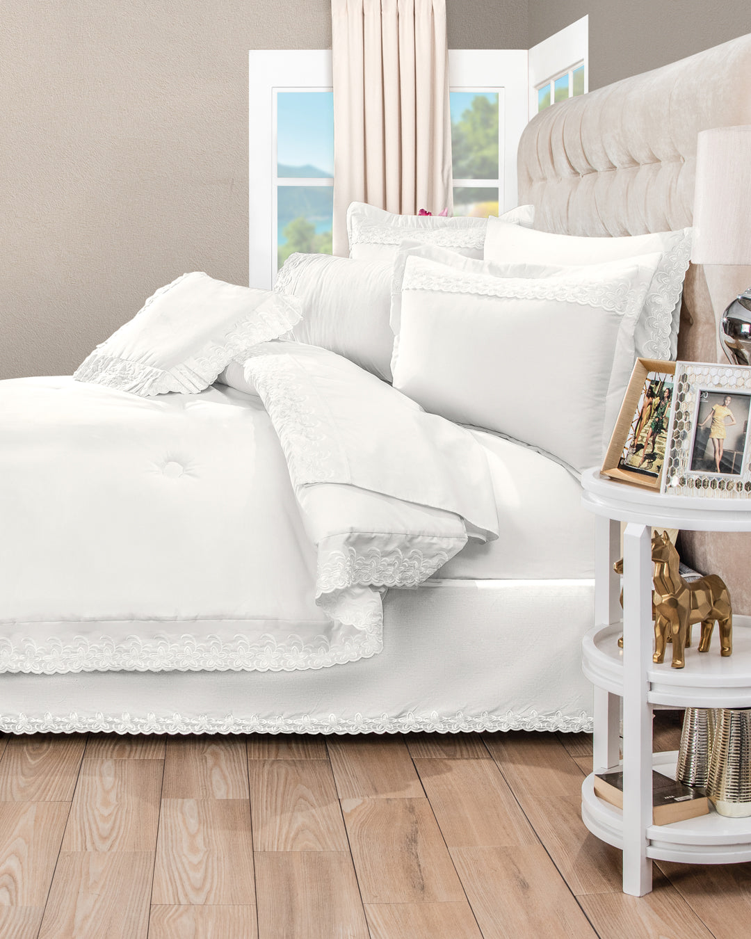 A neatly made bed with the Intima Hogar Juego de Edredón Bordado Fidanza, decorative shams, and lace-trimmed bedding sits by a round nightstand with photos, gold figurines, and a lamp, under beige curtains on light wood floors.