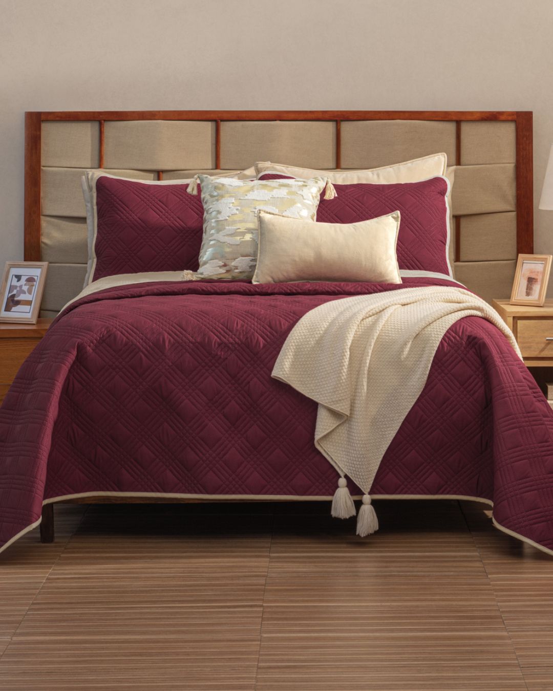 A neatly made bed features the Intima Hogar Juego de Edredón Ligero Burdeos with beige pillows and a tassel-accented throw. Wooden nightstands with lamps and framed photos flank the bed, all on a warm wooden floor.