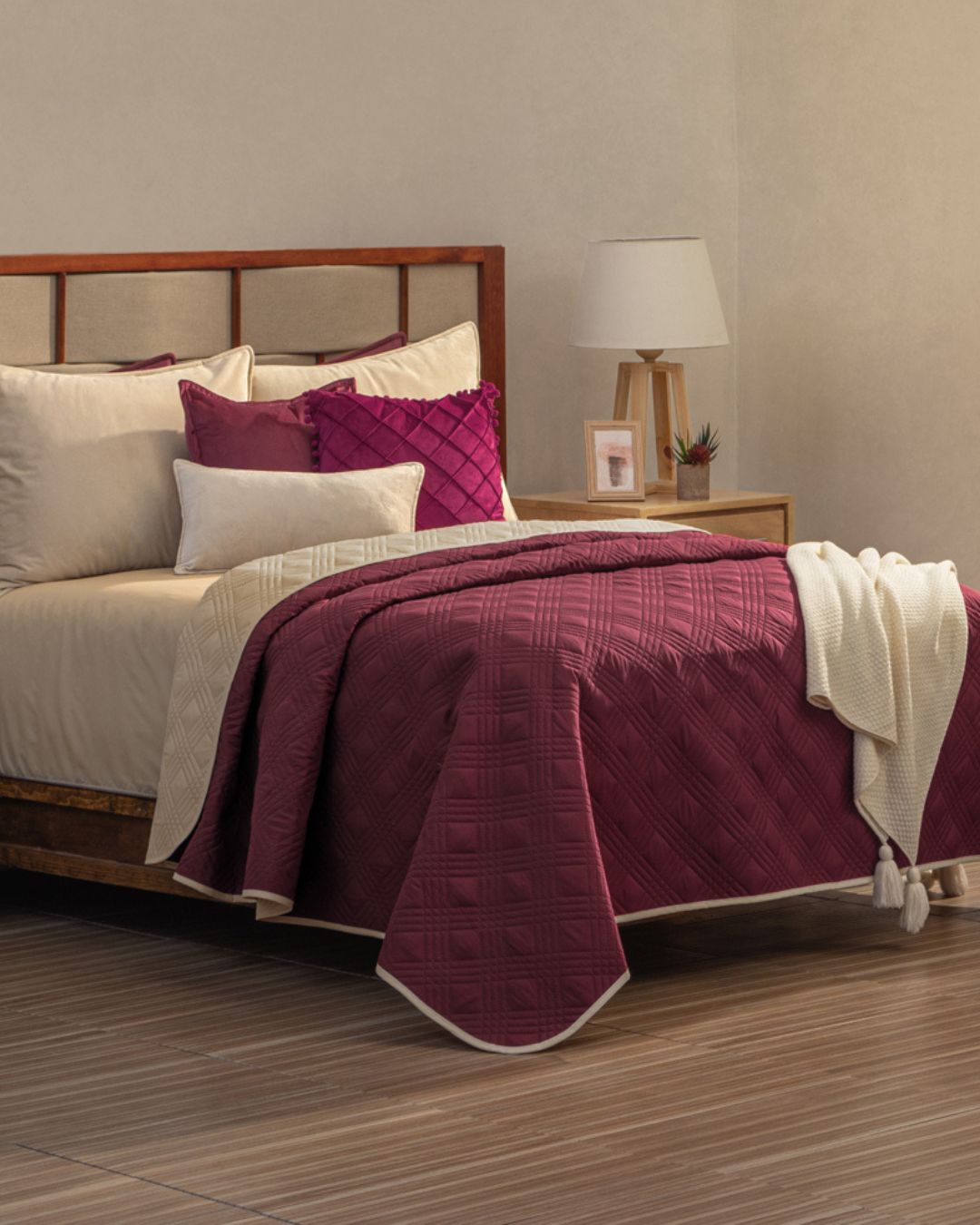 A neatly made bed showcases the Intima Hogar Juego de Edredón Ligero Burdeos, a reversible burgundy and cream bedding set with matching decorative pillow covers. Wooden floors and neutral walls complete the stylish room, accented by a cozy bedside table.