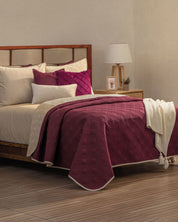 A neatly made bed showcases the Intima Hogar Juego de Edredón Ligero Burdeos, a reversible burgundy and cream bedding set with matching decorative pillow covers. Wooden floors and neutral walls complete the stylish room, accented by a cozy bedside table.