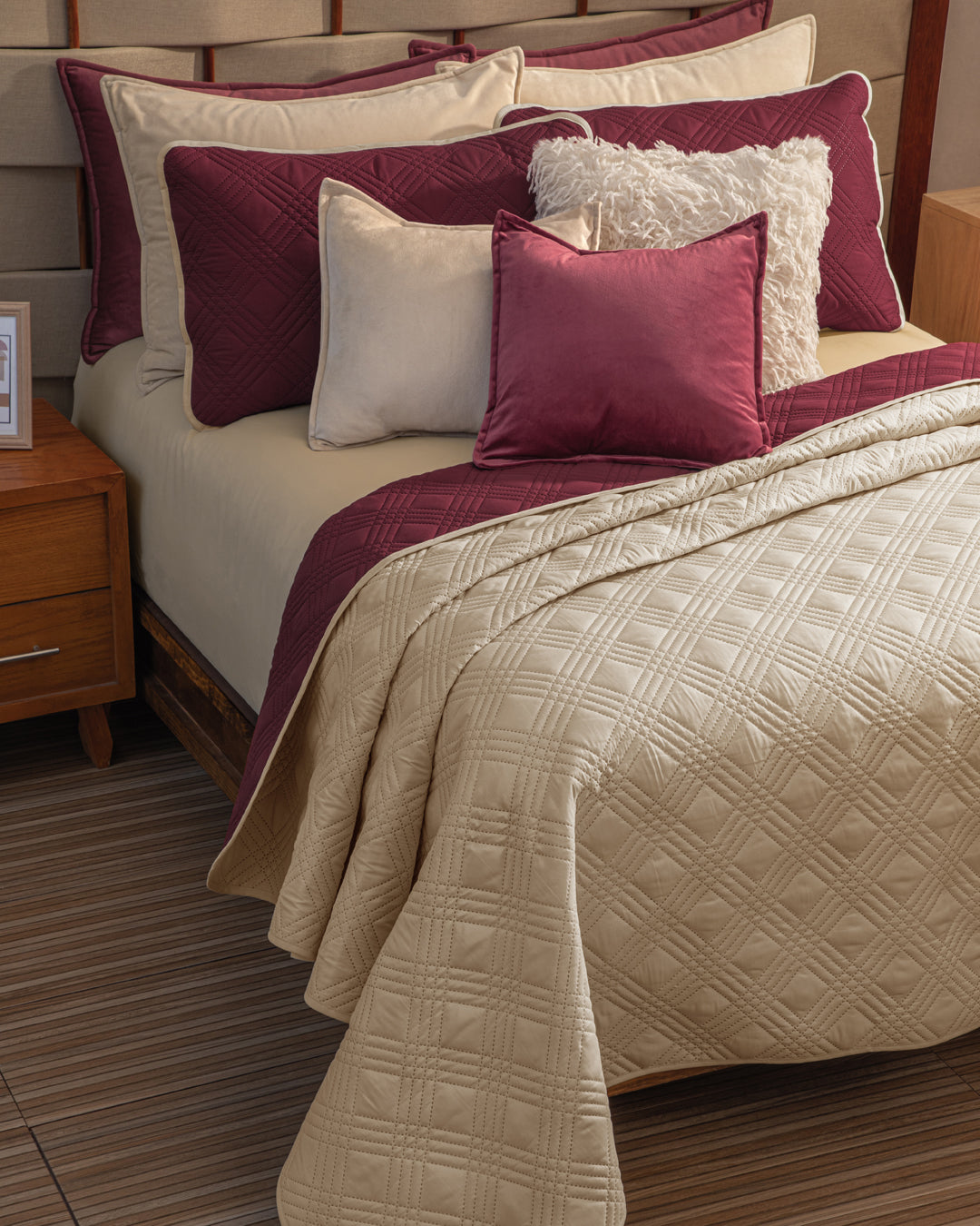 A cozy bedroom features the Juego de Edredón Ligero Burdeos by Intima Hogar, with cream quilted bedding, burgundy accent pillows, a fluffy white pillow, wooden furniture, and striped flooring.