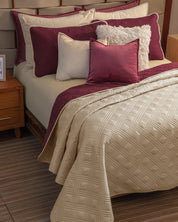 A cozy bedroom features the Juego de Edredón Ligero Burdeos by Intima Hogar, with cream quilted bedding, burgundy accent pillows, a fluffy white pillow, wooden furniture, and striped flooring.