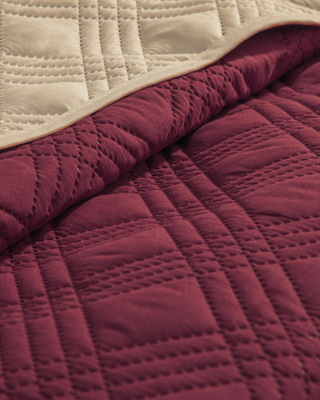 A close-up of Intima Hogar’s Juego de Edredón Ligero Burdeos shows its quilted burgundy side with stitched geometric patterns and smooth beige underside, partially folded to highlight both colors and textures.