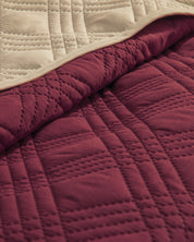 A close-up of Intima Hogar’s Juego de Edredón Ligero Burdeos shows its quilted burgundy side with stitched geometric patterns and smooth beige underside, partially folded to highlight both colors and textures.