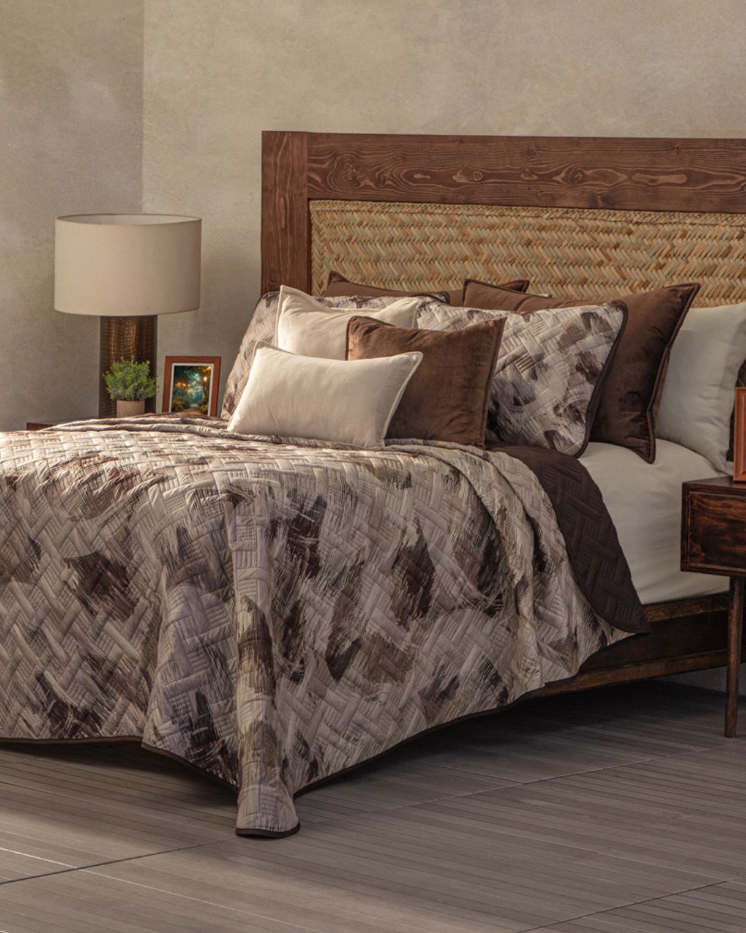 A cozy, elegant habitación features a wooden bed frame with the Juego de Edredón Ligero Coffee by Intima Hogar, accent pillows, a nightstand with lamp, small plant, and framed photo on a grey tiled floor and beige wall.