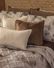 A neatly made bed displays the Intima Hogar Juego de Edredón Ligero Coffee, accented with beige and brown throw pillows—one rectangular, one square—arranged before matching patterned pillows.
